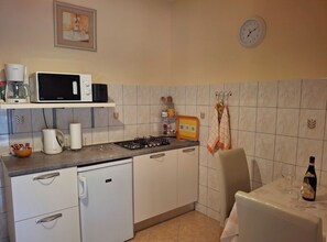 A compact kitchen with a microwave, toaster, kettle, and a small refrigerator. There is a clock on the wall and a table with a bottle and glasses. - Apartments Saulan (Konavle)