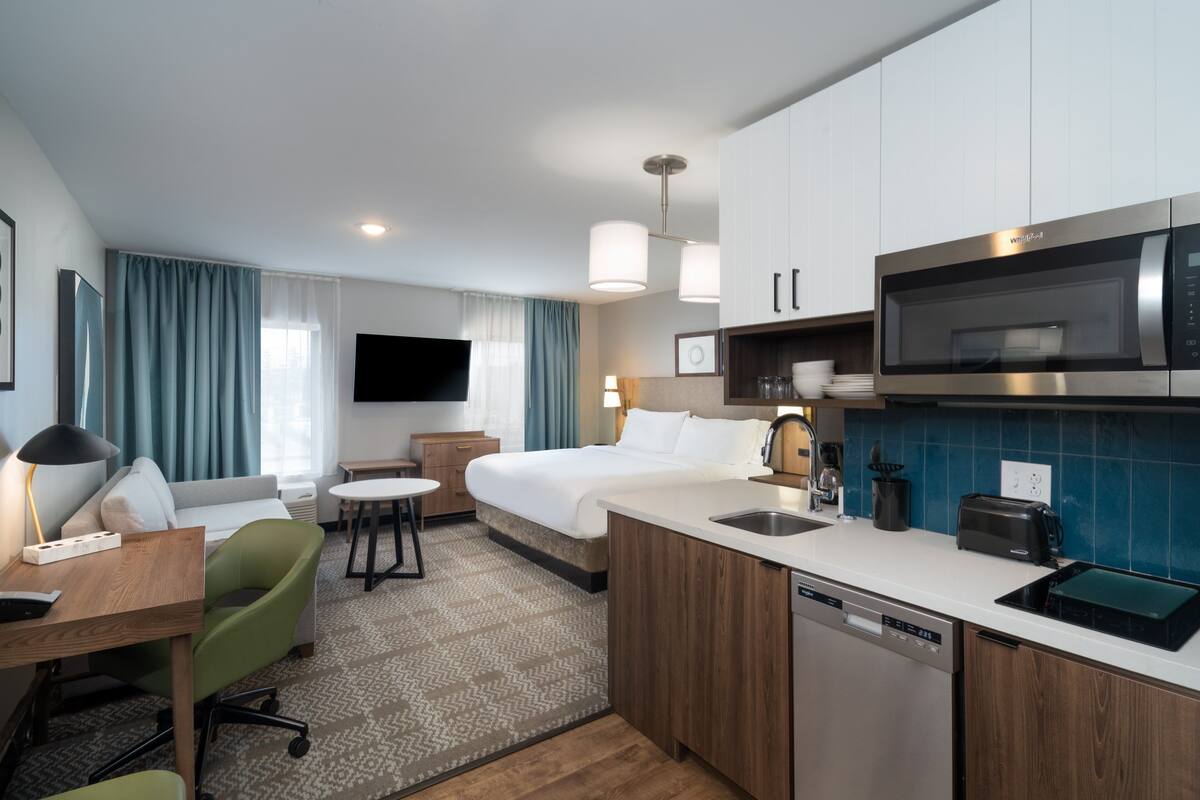 Habitación de hotel con cama, escritorio con silla, kitchenette con microondas y televisión.