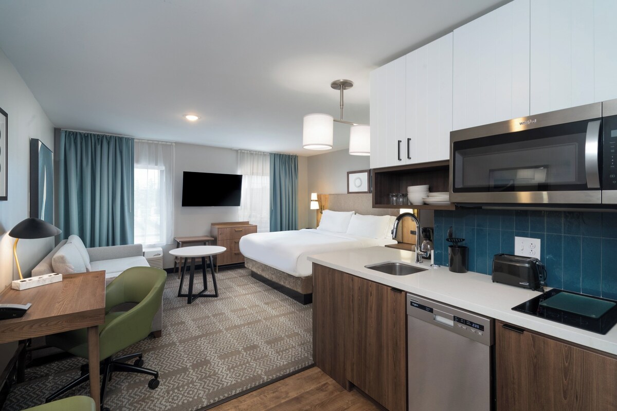 Habitación de hotel con cama, escritorio con silla, kitchenette con microondas y televisión.