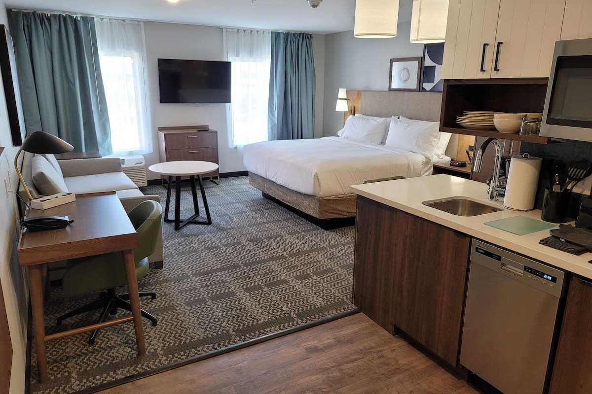 Habitación de hotel con una cama, un escritorio con silla, una mesita, un sofá, una zona de cocina con fregadero y lavavajillas, y un televisor de pantalla plana montado en la pared.