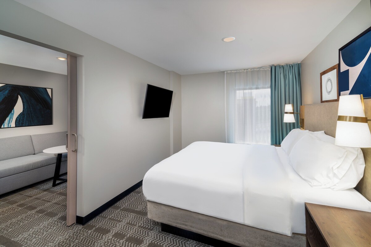 Una habitación de hotel con cama, sofá, televisión y ventana con cortinas.