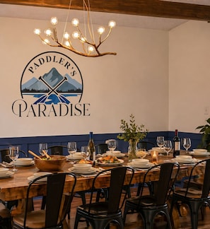 Un coin repas avec une table en bois dressée pour six personnes, un lustre et un logo de « Paddler’s Paradise » fixé au mur.