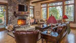 Un salón acogedor con chimenea de piedra, muebles de cuero y grandes ventanales con vistas a árboles.
