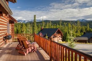 A wooden deck with lounge chairs and a barbecue grill, overlooking a forest and mountains.