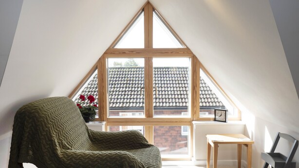 A cozy attic room with a sloped ceiling, a wooden chair, a small table, and a view of a tiled roof through a large window. - Adelaide Place (Canterbury)