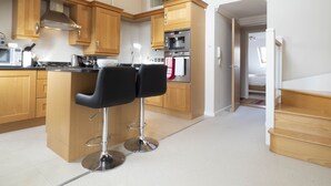 A modern kitchen with wooden cabinets, a central island with two black bar stools, a stainless steel oven, and a microwave. - Adelaide Place (Canterbury)