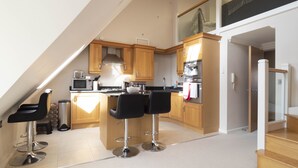 A modern kitchen with wooden cabinets, a central island with bar stools, a microwave, and a built-in oven. - Adelaide Place (Canterbury)