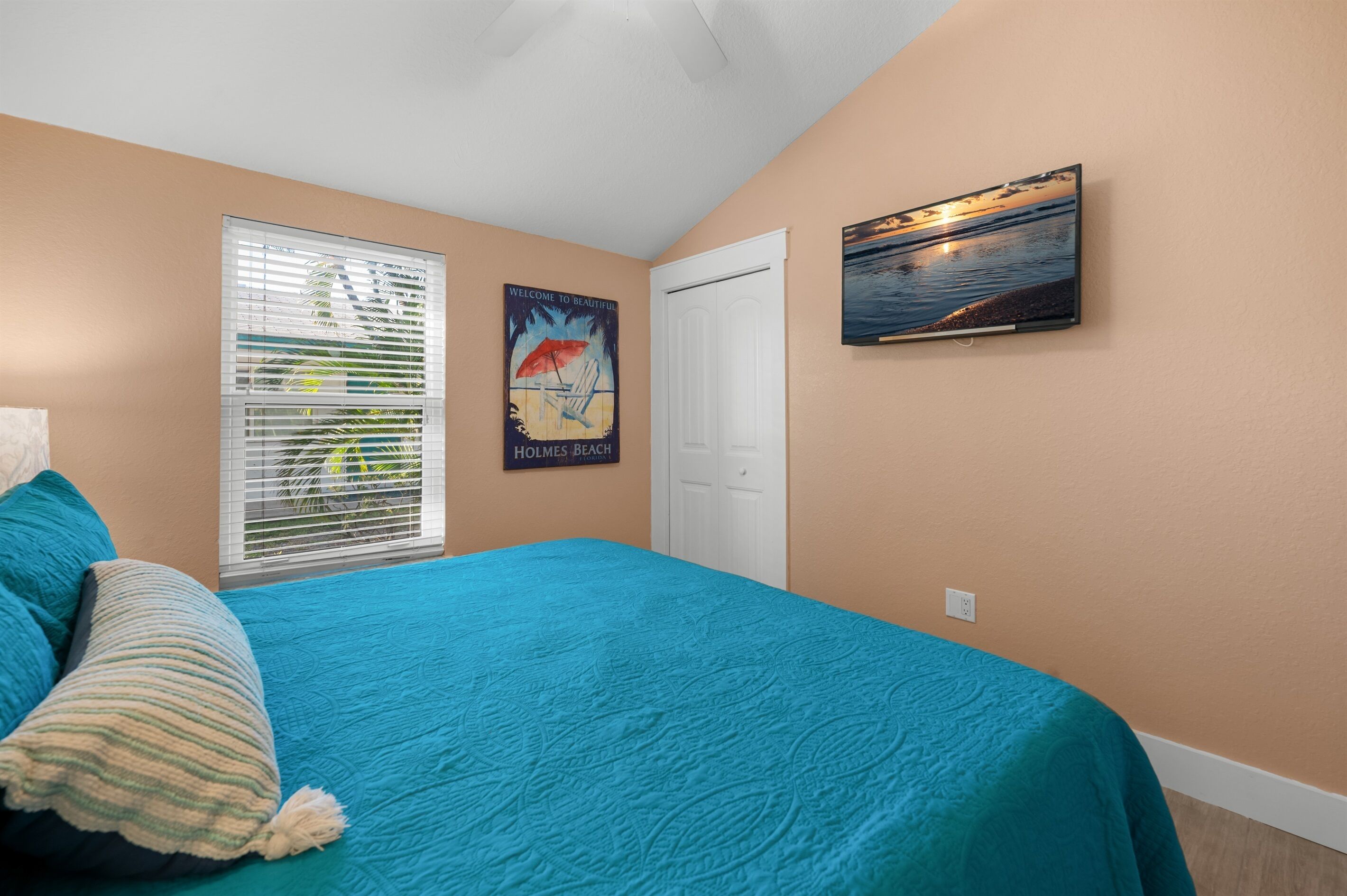 Un dormitorio con colcha color turquesa, un cartel con motivos playeros, un televisor en la pared y una puerta blanca.