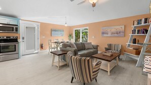 Uma sala de estar moderna com sofá, poltrona listrada e mesa de jantar. Há uma cozinha com micro-ondas e forno.