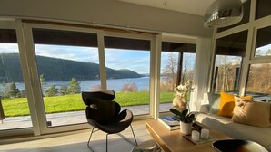 A modern living room with a large sliding glass door leading to a scenic view of a lake and mountains. - Cabin Oslofjord -sleeps14-seaview-familyfriendly (Frogn)