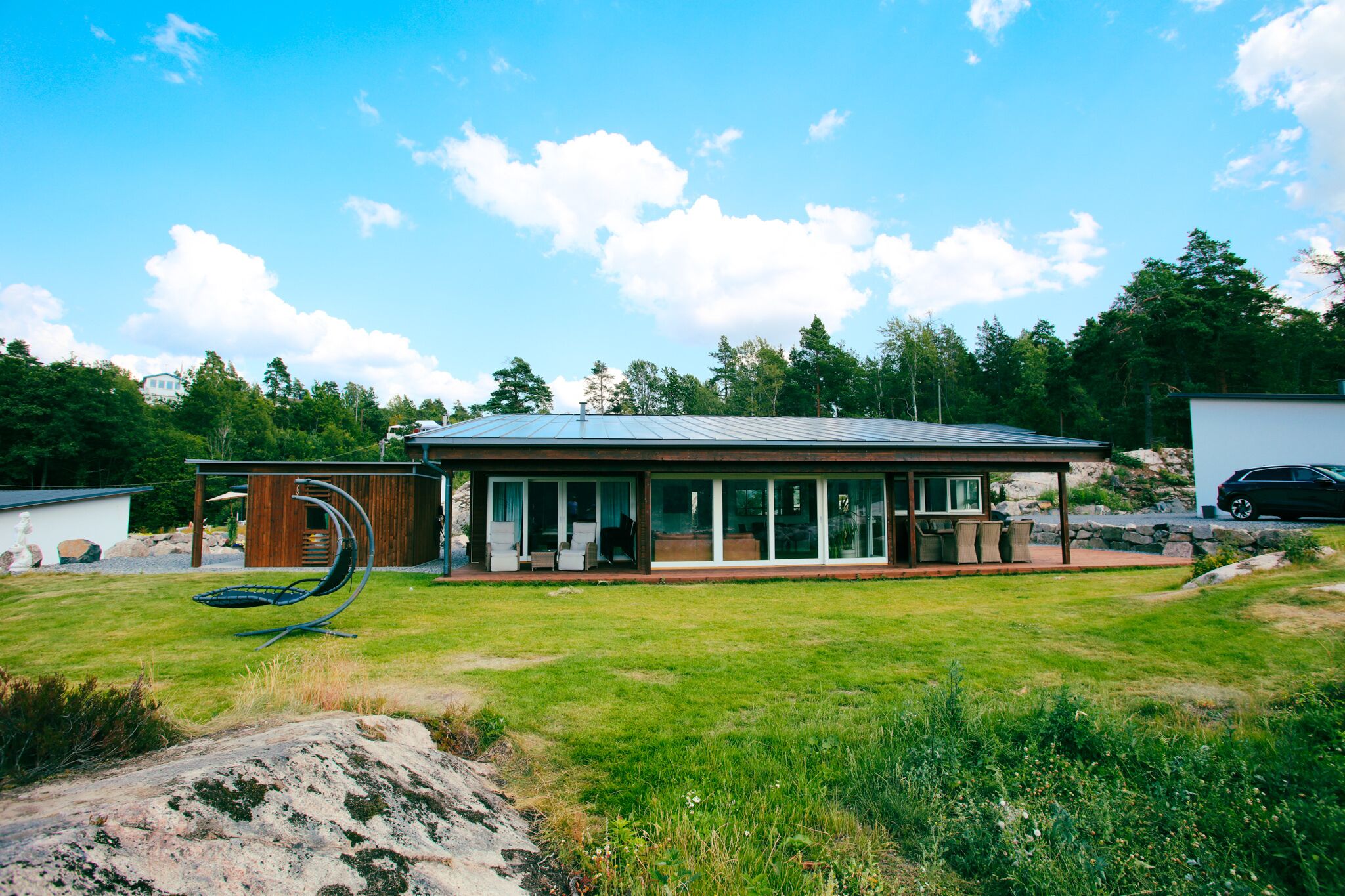 Una casa moderna con un amplio ventanal, un patio con mobiliario exterior y un área verde circundante.