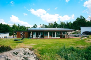 Une maison moderne dotée d’une grande façade vitrée, d’un patio avec mobilier d’extérieur et d’une pelouse verdoyante tout autour.