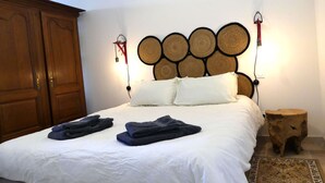 Ein ordentlich gemachtes Bett mit weißen Leinentüchern und zwei gefalteten Handtüchern, ein Holzschrank und ein rundes hölzernes Kopfteil mit gewebten Paneelen.
