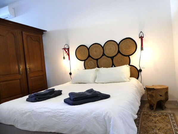 Un lit bien fait, avec du linge de lit blanc et deux serviettes pliées, une armoire en bois et une tête de lit ronde en bois ornée de panneaux tissés.
