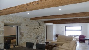Ein geräumiger Wohnbereich mit Steinwand, Holzbalken, einem Esstisch mit Stühlen, einer Couch und einem Fenster, das natürliches Licht hereinlässt.