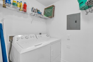 A laundry room with a front-loading washer and dryer, a shelf with cleaning supplies, and a power outlet on the wall.