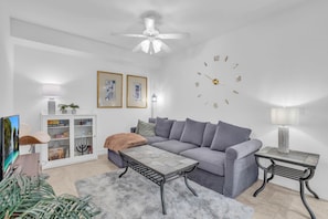 Un salon moderne avec un canapé gris, une table basse, une table d’appoint, un ventilateur de plafond et une horloge murale.