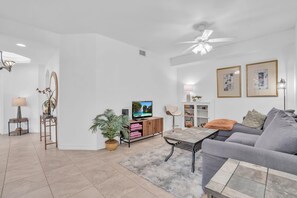 A modern living room with a grey sofa, a coffee table, a TV on a wooden cabinet, a ceiling fan, and a potted plant. - Beautiful Corner Unit in Woods Edge W/ Brand New Kitchen, Community Pool -Close to Beach (Bonita Springs)