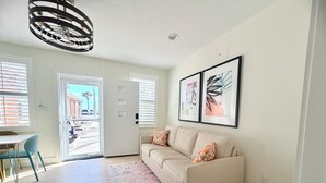 Ein Wohnzimmer mit einem beigefarbenen Sofa, einem gemusterten Teppich, einem Essbereich mit Tisch und Stühlen sowie Kunstwerken an der Wand.