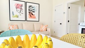 Ein Zimmer mit einem weißen Tisch, einem blauen Stuhl, einer beigen Couch mit gemusterten Kissen und zwei gerahmten Kunstwerken an der Wand.