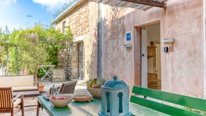 Un espace extérieur aménagé avec une table en bois, des chaises et un banc vert. On y trouve également un mur en pierre et une porte donnant accès à l’intérieur.
