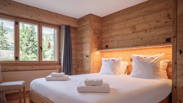 A wooden cabin room with a bed, two windows, a chair, and a view of trees.