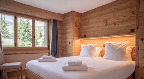 A wooden cabin room with a bed, two windows, a chair, and a view of trees.