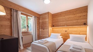 A wooden cabin room with two beds, a desk, and a window with curtains.