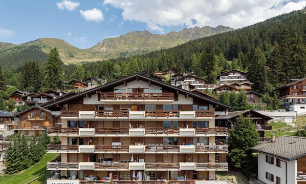 A multi-story hotel with balconies, set in a mountainous area with dense forest.