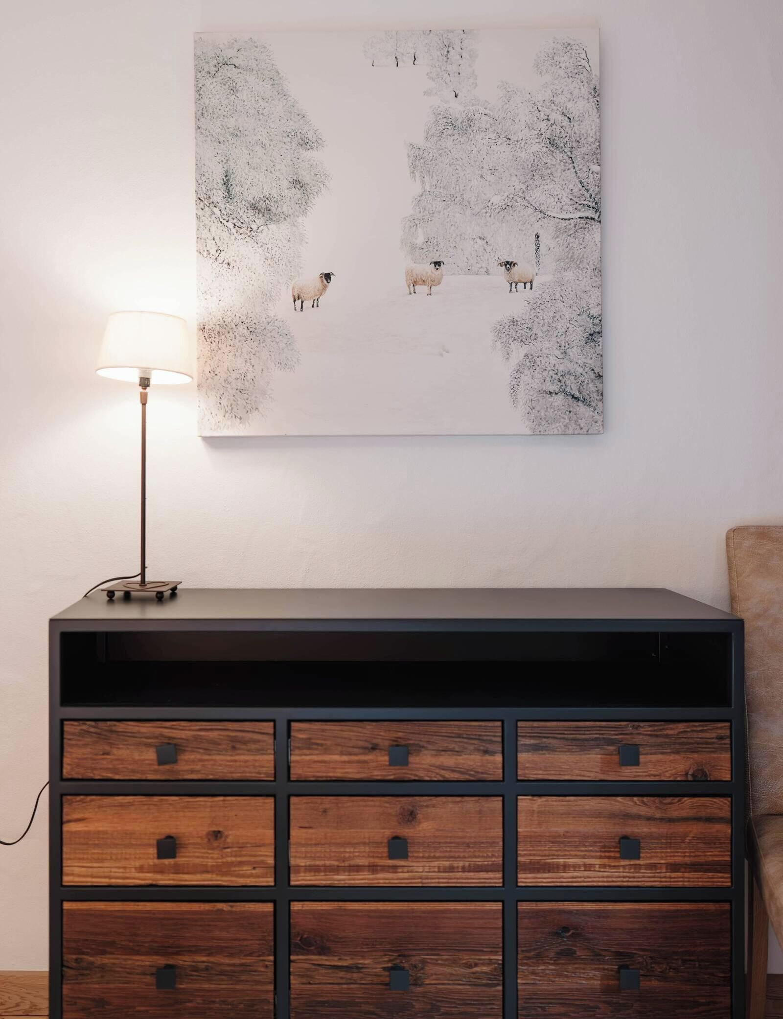 A room with a wall-mounted abstract painting, a table with a lamp, and a wooden cabinet with drawers.