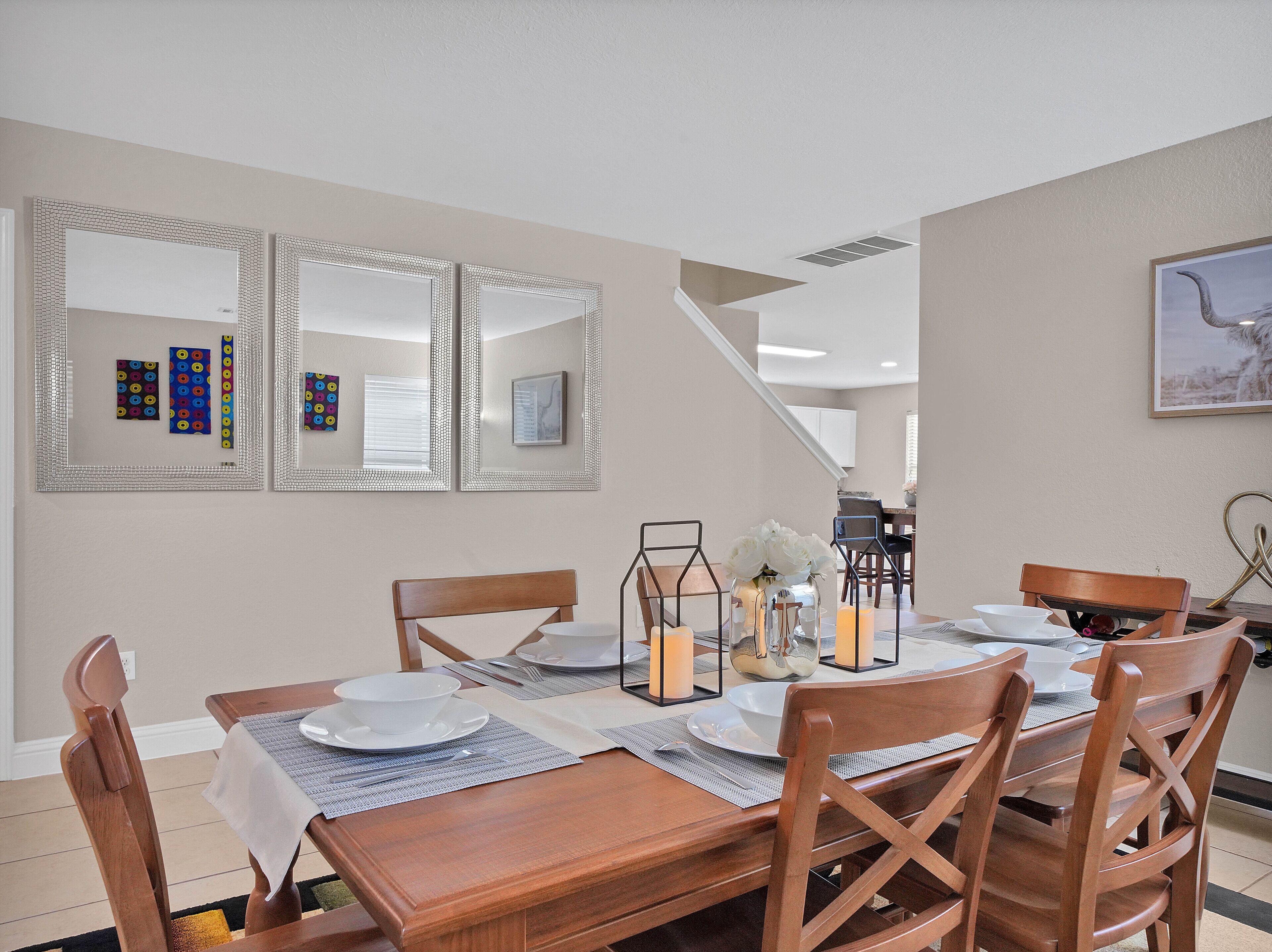 A dining area with a wooden table set for four, surrounded by chairs. There are three mirrors on the wall, and a painting above the table.