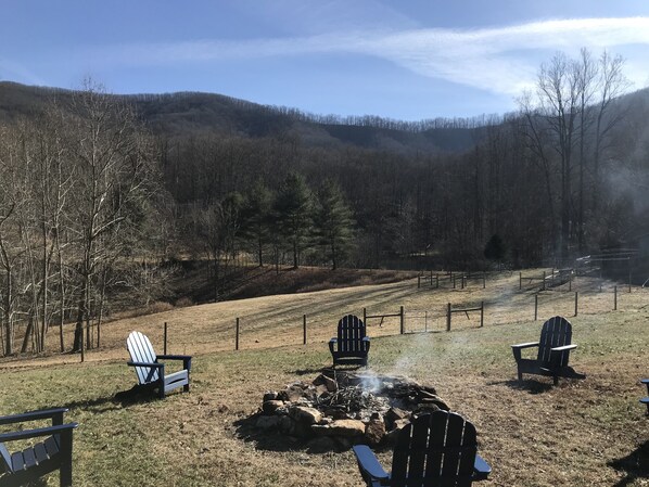 A campsite with Adirondack chairs, a fire pit, and a fenced area with a view of a forested hillside.
