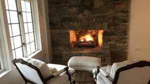 Une cheminée en pierre avec un feu, deux fauteuils blancs rembourrés et une petite table ronde dans une pièce dotée d’une fenêtre.