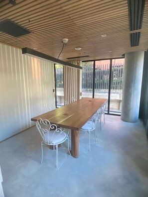 A spacious room with a long wooden table and white metal chairs, large windows with a view of a fence, and a ceiling with a wooden pattern. - 23 Hotel (Medellín)