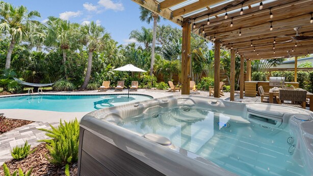 Un jacuzzi y una piscina con sillones de descanso y sombrillas, rodeado de palmeras y una pérgola de madera.