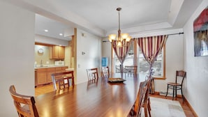 Un comedor amplio con una mesa y sillas de madera, una cocina al fondo y un ventanal con cortinas.