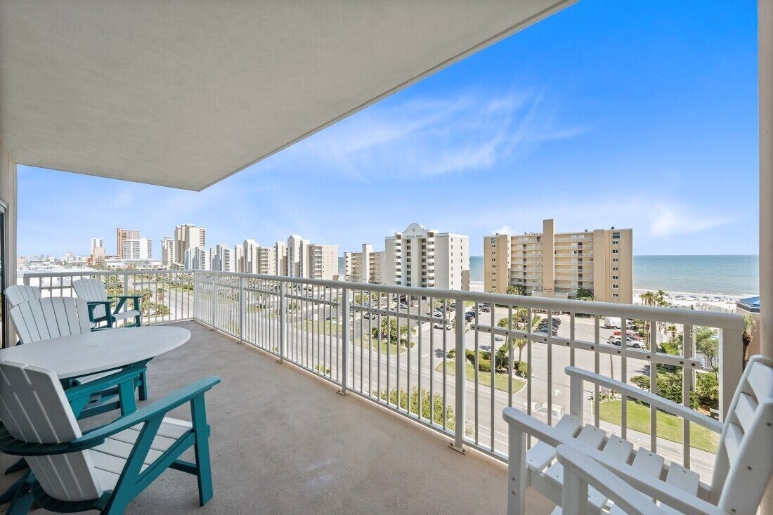 A balcony with a table and chairs, overlooking a cityscape and beach.