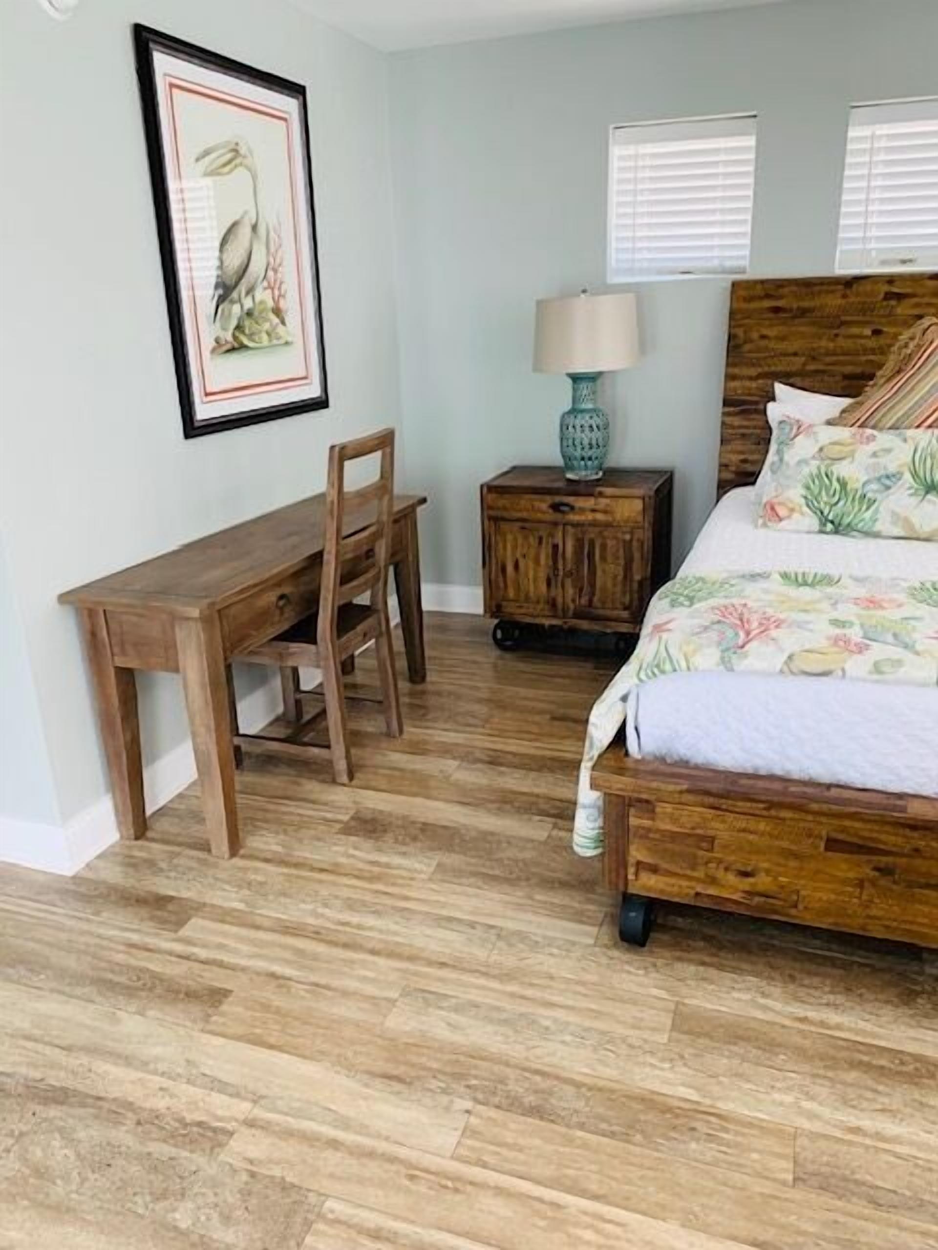 A bedroom with a wooden bed, a wooden desk, a chair, a nightstand with a lamp, and a framed picture on the wall.