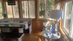 L’intérieur chaleureux d’un chalet avec une table à manger en bois, des bancs, une petite table ronde avec une lampe et de grandes fenêtres donnant sur la forêt.