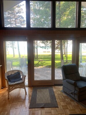 A room with a large sliding glass door leading to an outdoor deck, a wicker chair, a cushioned chair, and a woven rug.