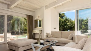 Un salon moderne avec une table basse en verre, un canapé beige et une vue sur une piscine et des arbres, visible à travers de grandes fenêtres.