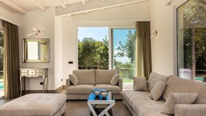 Un salon moderne avec un canapé, un pouf et une table basse. Il y a une fenêtre avec des rideaux et une vue sur les arbres.