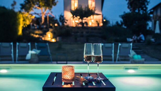 Ein Tisch am Pool mit zwei Weingläsern und einer Kerze, vor einem beleuchteten Gebäude im Hintergrund.
