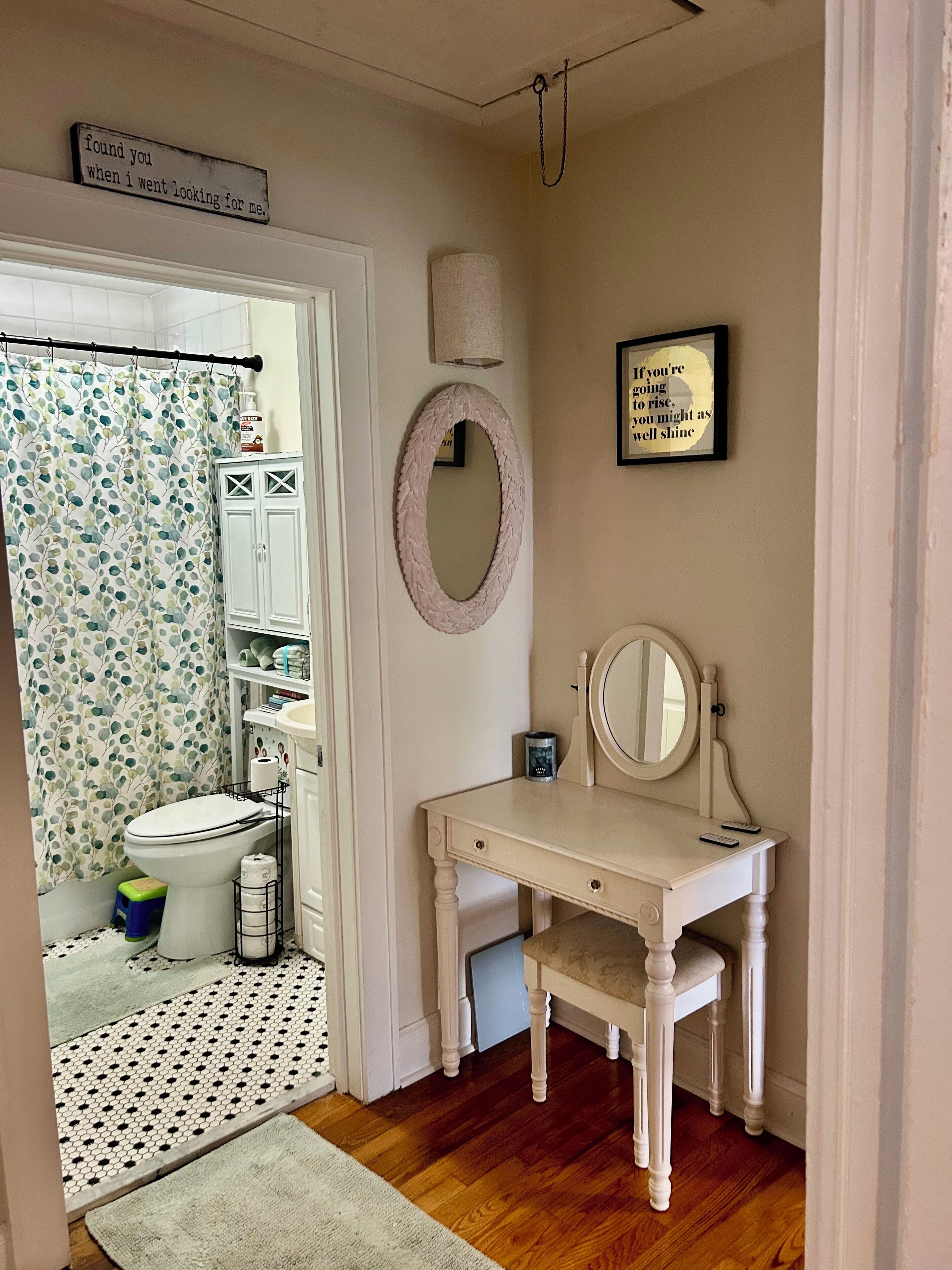 Une salle de bain équipée de toilettes, d’un lavabo et d’un meuble-lavabo avec un miroir. On y trouve un rideau de douche orné d’un motif de feuilles, un porte-serviettes et une citation encadrée accrochée au mur.