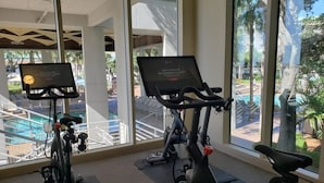 Un gimnasio interior con dos bicicletas estáticas, ventanas amplias y vistas a una piscina y palmeras.