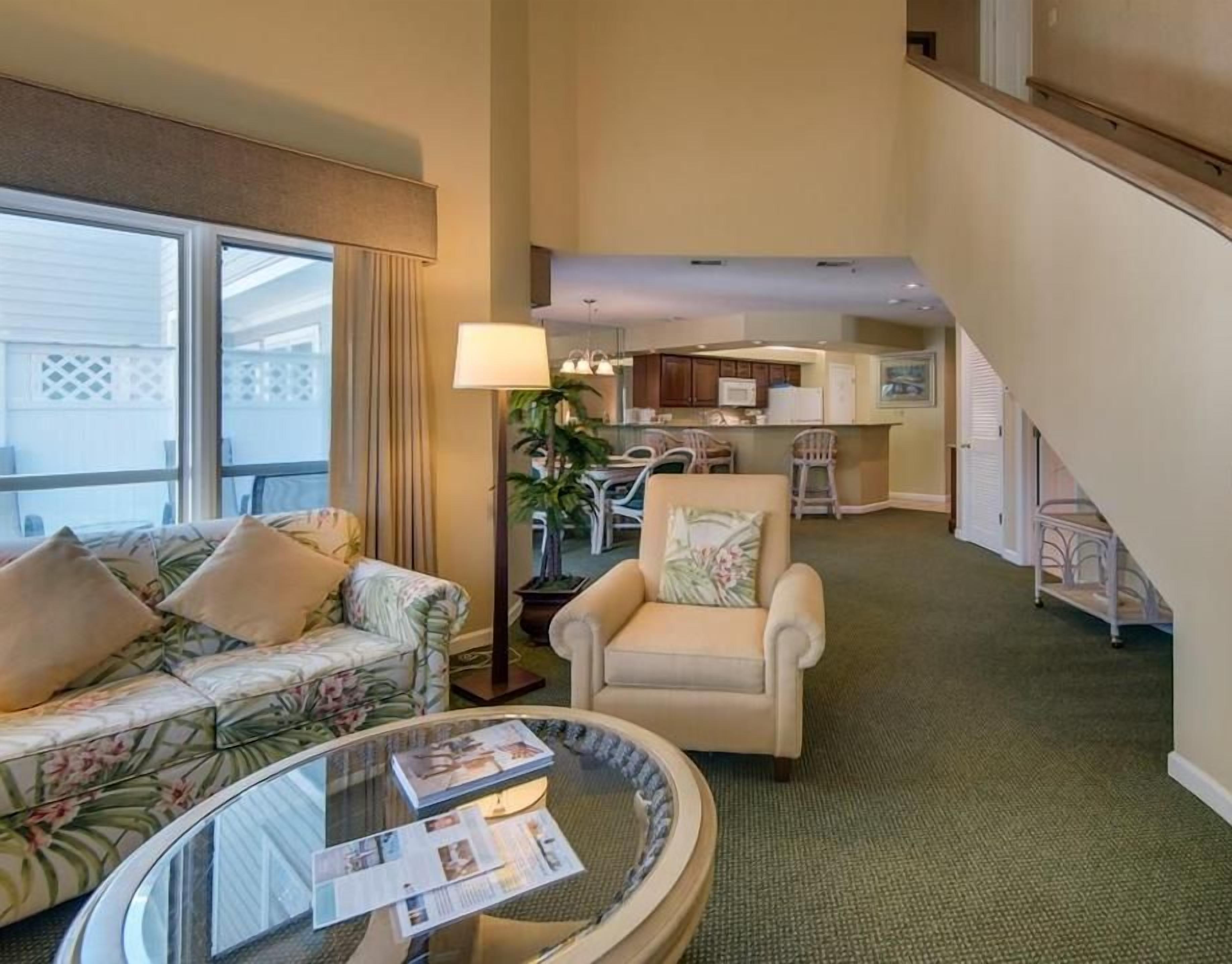 A spacious living area with a sofa, armchair, and a glass coffee table. A staircase leads to an upper level with a kitchen area visible.