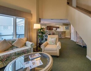 A spacious living area with a sofa, armchair, and a glass coffee table. A staircase leads to an upper level with a kitchen area visible.