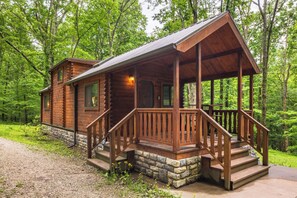 A log cabin with a porch, stone foundation, and a forested backdrop.
