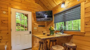 L’intérieur d’un chalet en bois, avec un téléviseur fixé au-dessus d’une table en bois, des tabourets et une porte dotée d’une fenêtre.