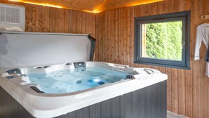 En stuga byggd av trä med ett bubbelbad, ett fönster med utsikt över grönskan och en handduk som hänger på väggen.